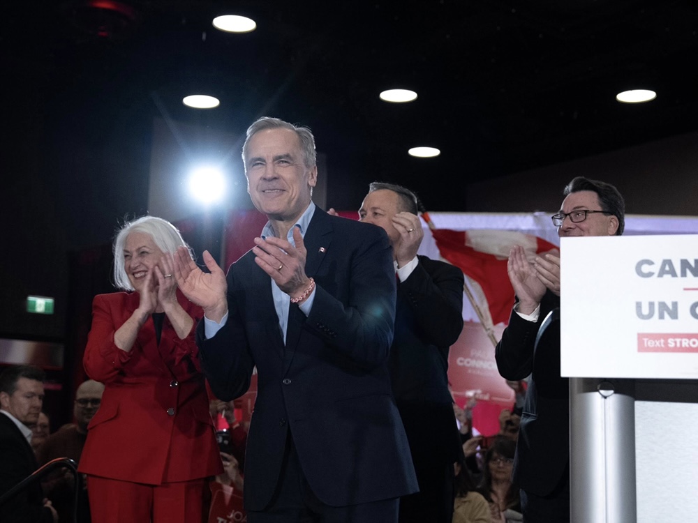 Mark Carney stands next to a podium, wearing a suit and smiling and clapping.