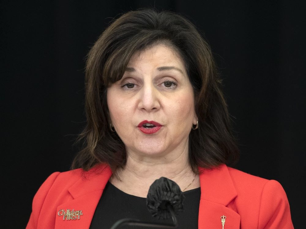 A middle-aged woman with light skin and dark hair, wearing a red blazer over a black blouse, speaks into a microphone.