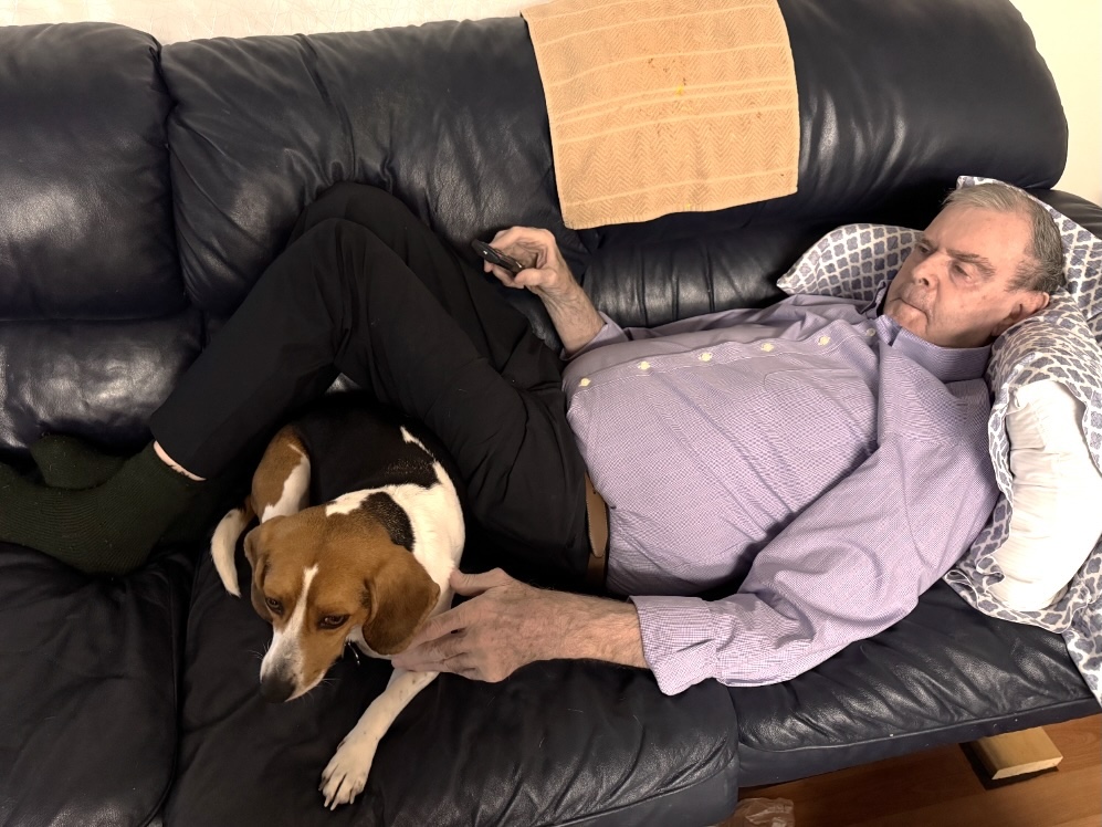 A man in his 80s with light skin and grey hair lies on a black couch, his head propped up by pillows. A beagle-like dog lies under his bent knees.