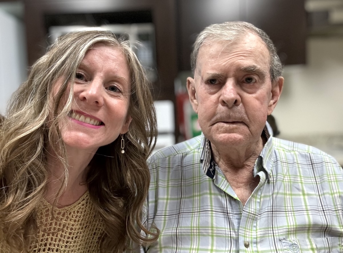 A middle-aged woman with light skin and long, wavy blond hair stands next to her father, a man in his 80s with light skin and short grey hair.