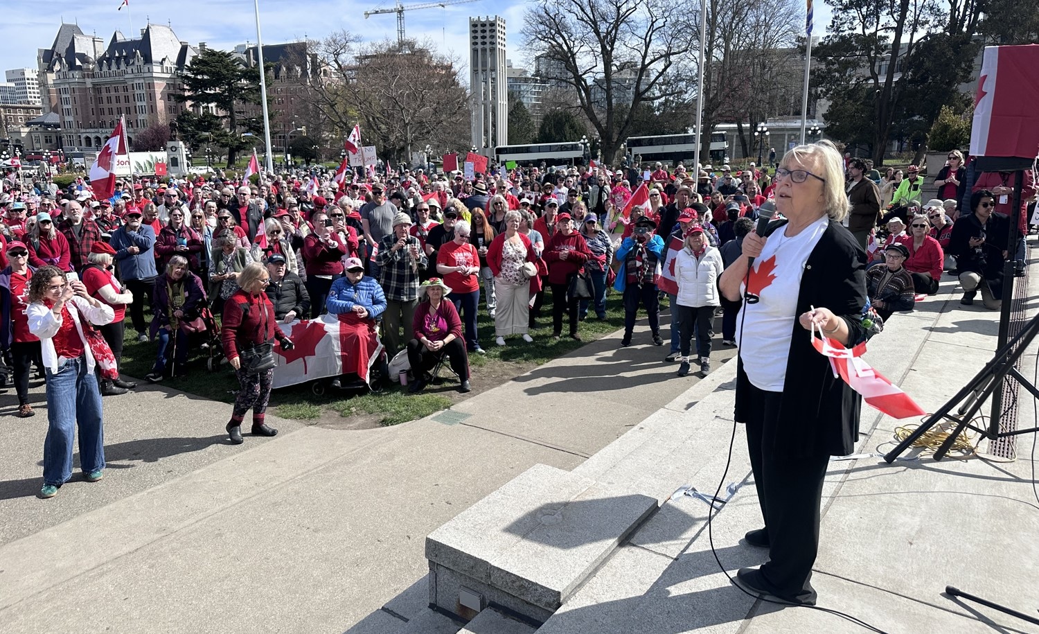 A 70-ish woman with light skin and blond hair, holding a Canadian flag in one hand and wearing a maple leaf T-shirt, stands on an outdoor stage addressing a crowd of people, many dressed in red, some holding Canadian flags.
