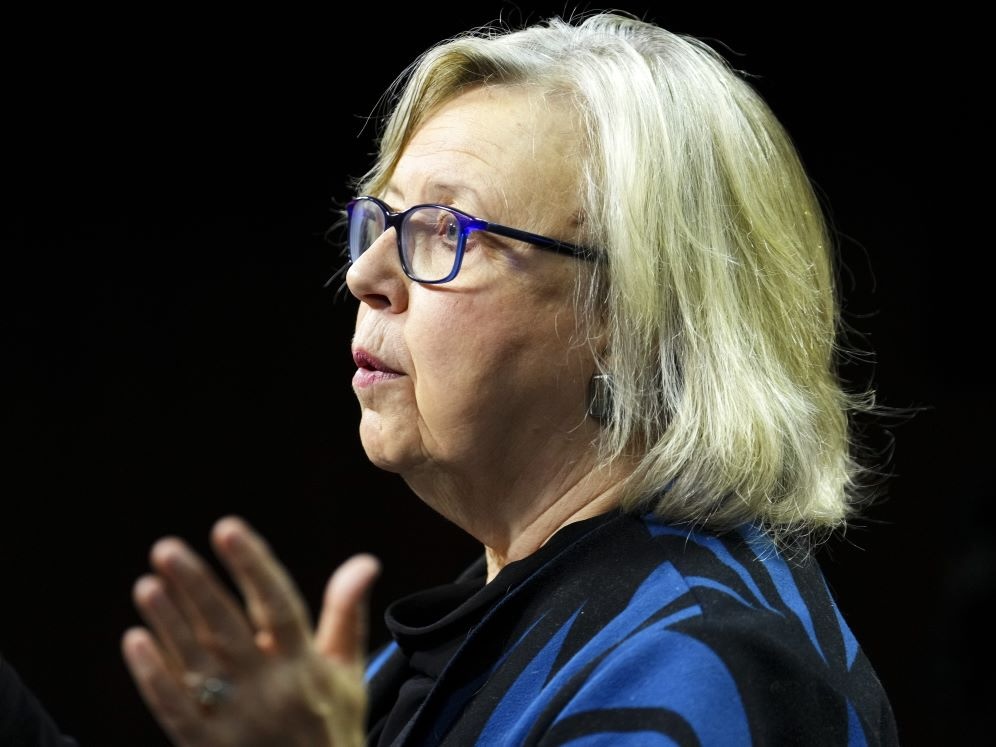 Profile of a 70-ish woman with light skin and blond hair, wearing thick-framed glasses and a dark blue blouse.