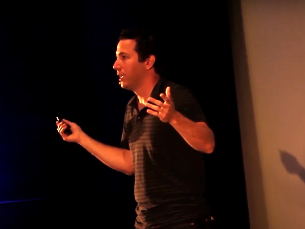 Orange light tinges an image of a main with short dark hair wearing a dark short-sleeved polo shirt. He is mid-speech, holding his arms out to the side.