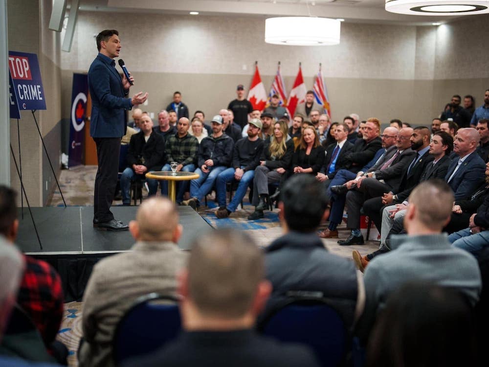 Poilievre is onstage with a ‘Stop the Crime’ sign behind him. Men and women in suits and jeans sit in chairs arranged in a semicircle in front of him.
