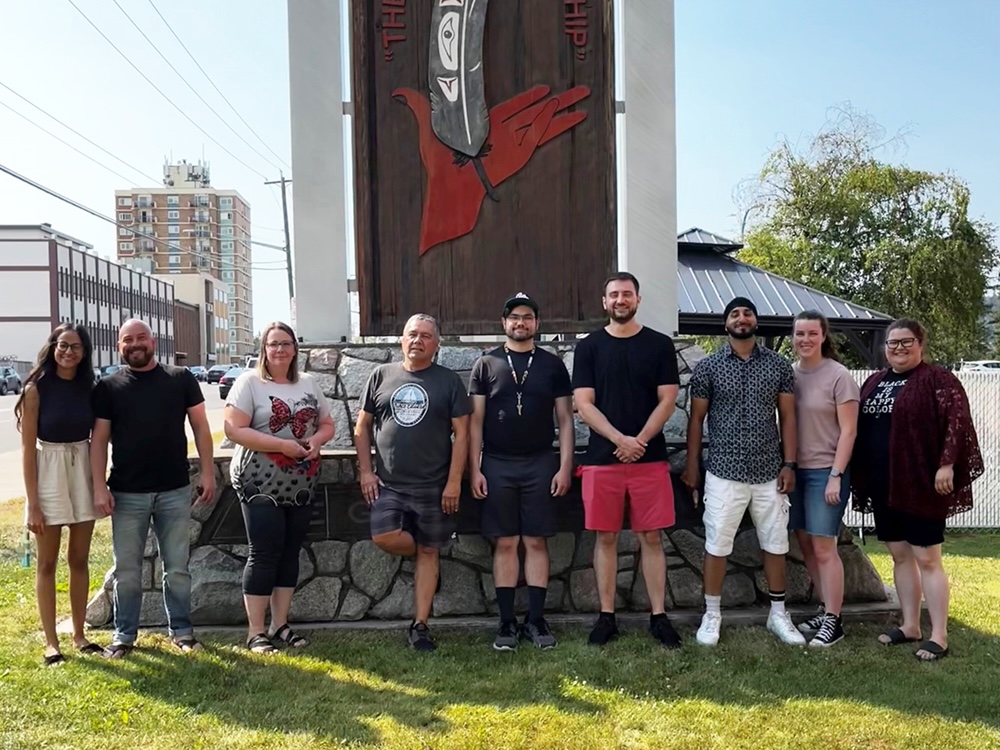 Nine people dressed casually stand on the grass in front of a large wooden sign for the Prince George Native Friendship Centre.