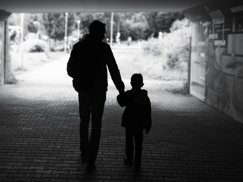 A black and white photo shows, from behind, a man and young boy, silhouetted, walking through a tunnel towards daylight.