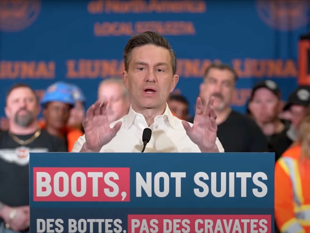 Pierre Poilievre stands at a blue and red lectern that reads ‘Boots, Not Suits.’ He is wearing a white button-down shirt and has brown slicked-back hair and pale skin.