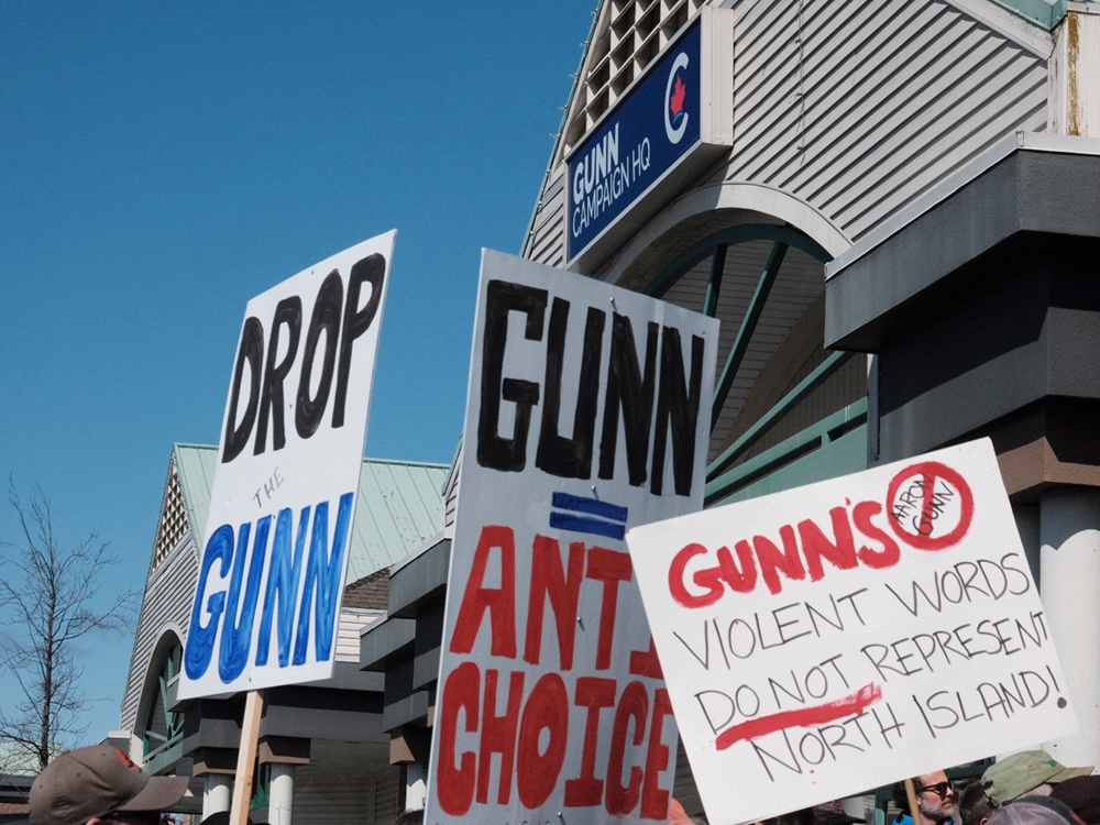 A sign on a building reads, ‘Gunn campaign HQ.’ People are gathered holding homemade signs. They read, ‘Drop the Gunn,’ ‘Gunn = Anti-Choice’ and ‘Gunn’s violent words do not represent North Island.'