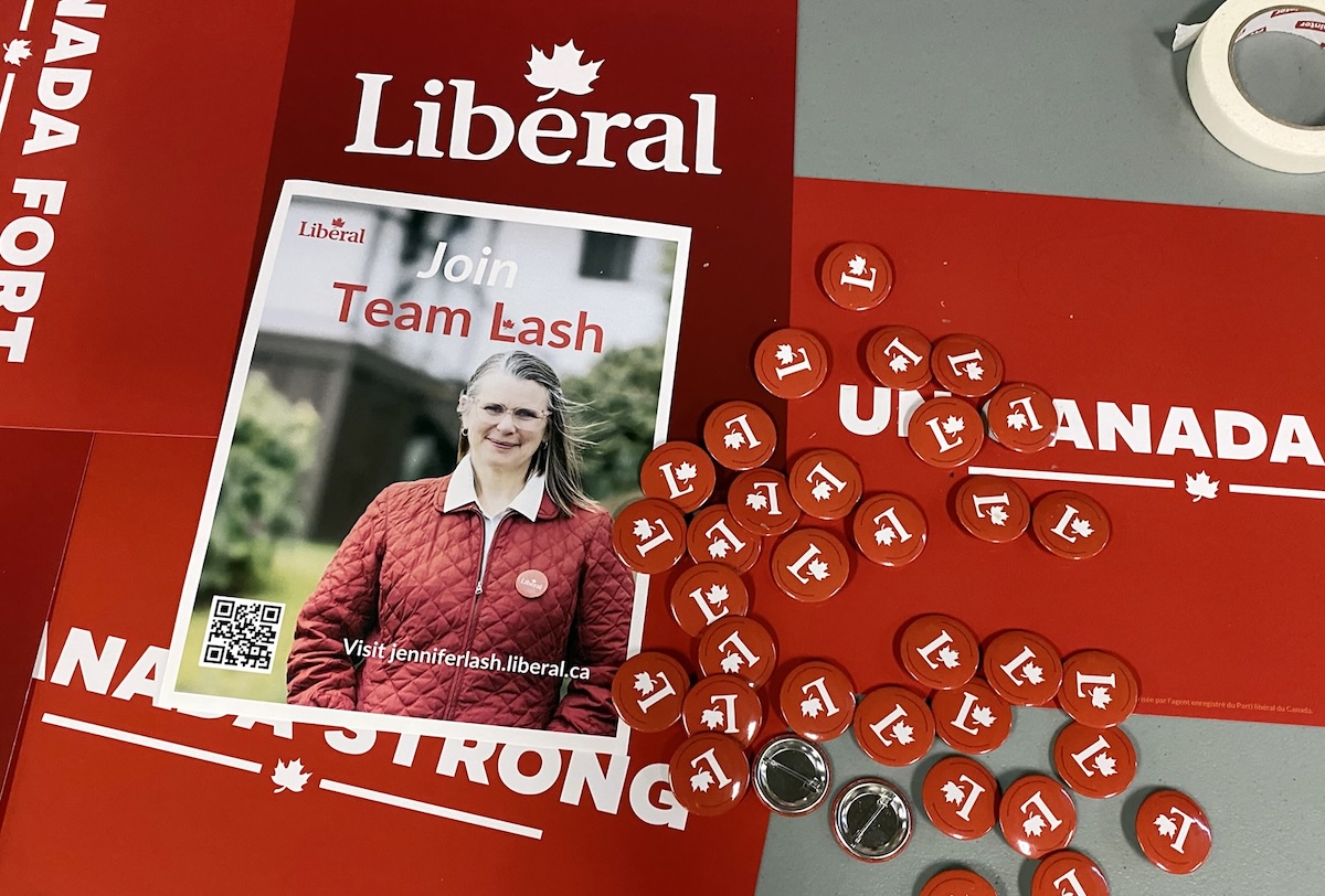 A poster reads, ‘Join Team Lash.’ Red pins with Liberal L’s are scattered on the table.
