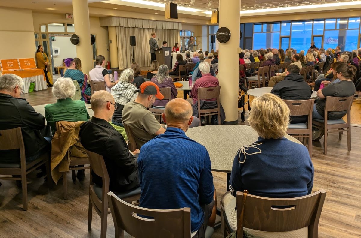 A room full of people sitting on chairs in rows and around tables, with four people at the front speaking into microphones.