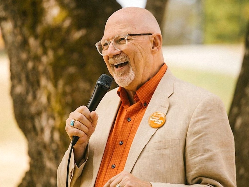 A bald man with a grey and white goatee and clear-framed glasses speaks into a microphone outdoors. He is wearing a beige jacket over an red-orange shirt, with an orange pin on his lapel that says ‘Re-elect Randall Garrison, NDP.’