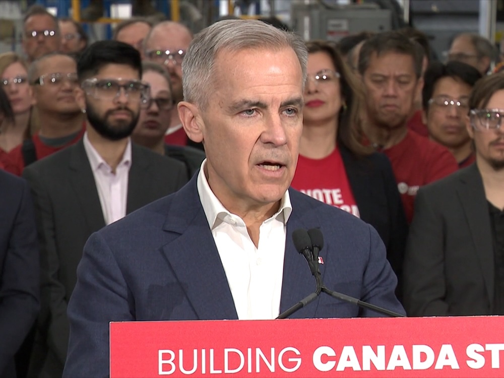 A man wearing a blue suit jacket and white button-down shirt stands at a lectern that bears a red sign that reads ‘Building Canada strong.’