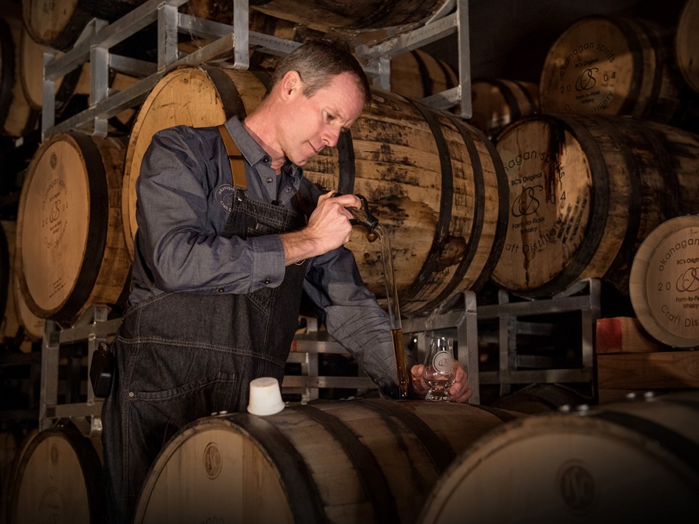 A man with short, dark hair wearing overalls and a button-up shirt pours liquid into a glass. Behind him are rows of wooden barrels.
