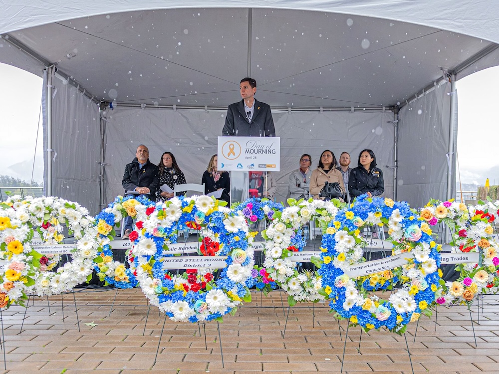 A man with short brown hair and pale skin tone stands at a lectern with a white sign that reads &lsquo;Day of Mourning.&rsquo; He is wearing a button-down shirt under a long grey jacket. People are seated onstage behind him under a white tent. Colourful memorial floral wreaths are arranged on a beige tile floor in front of the stage.