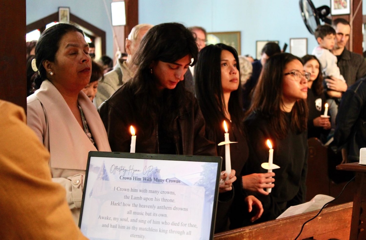 Mourners fill a church. In the foreground, four women stand in a pew, three of them holding lit candles. A computer screen shows the words to 'Crown Him with Many Crowns.'