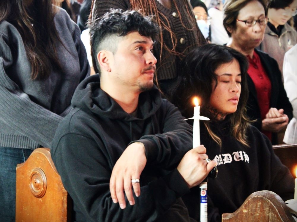 A man and woman sit in a pew in a church, with people standing in pews behind them. The man has medium-light skin and dark har and is holding a lit candle. The woman has medium-light skin and wavy long hair and has her eyes closed.