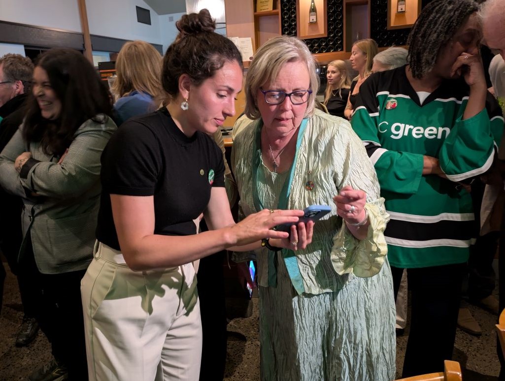 A light skinned older woman with grey hair, dark framed glasses and a light green dress peers at a phone held by a younger person with hair in a bun and in a black shirt and light pants. They are both surrounded by a crowd of people.