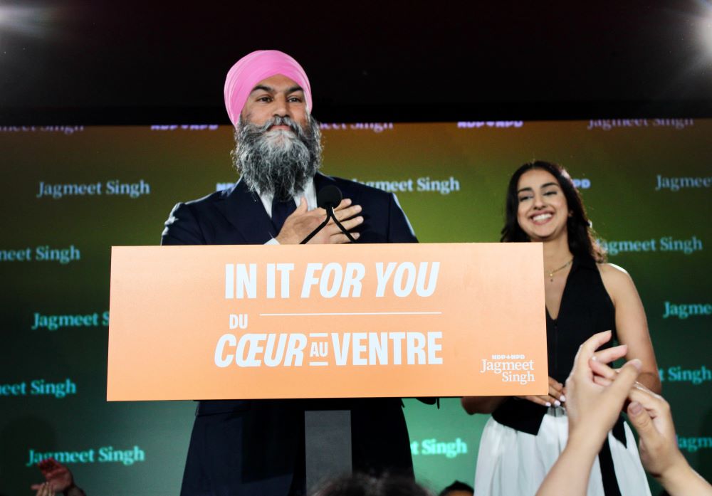 A bearded medium skin toned man with a pink head wrap and a suoit holds his hand to his heart as he speaks from a podium with a sign that reads “In It For You.” A medium skin toned woman in a black blouse and white skirt smiles beside him.