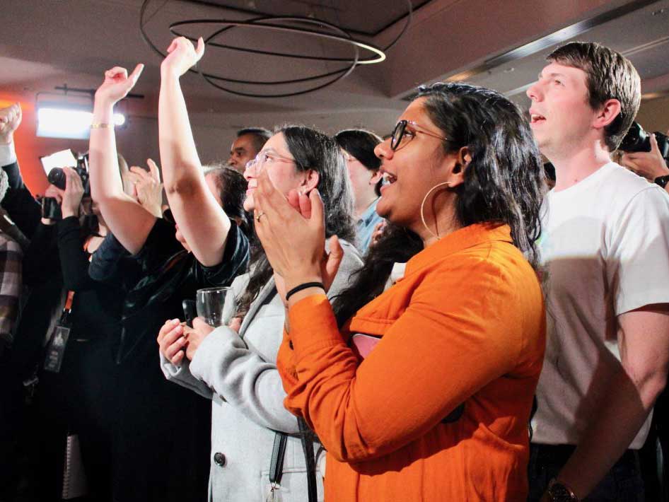 A medium-dark-skin-toned woman with long dark brown hair and a bright orange jacket on claps and smiles facing the leftmost side of the photograph. Behind her, a crowd of more cheering supporters.