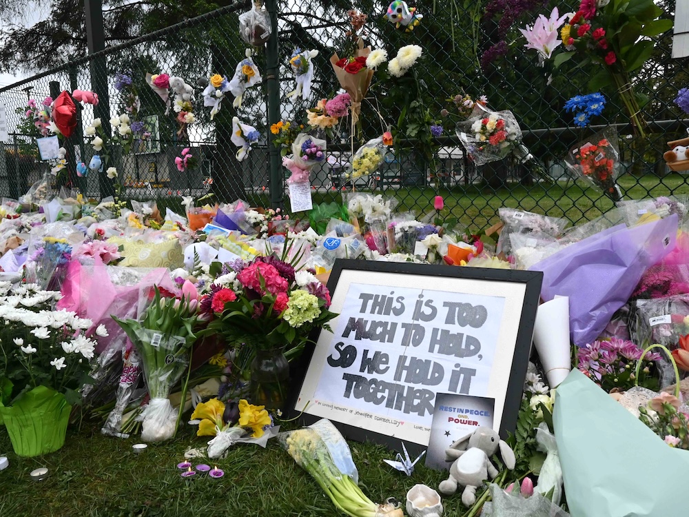 Flowers and a sign that says ‘This is too much to hold, so we hold it together’ are placed in front of a chain-link fence.
