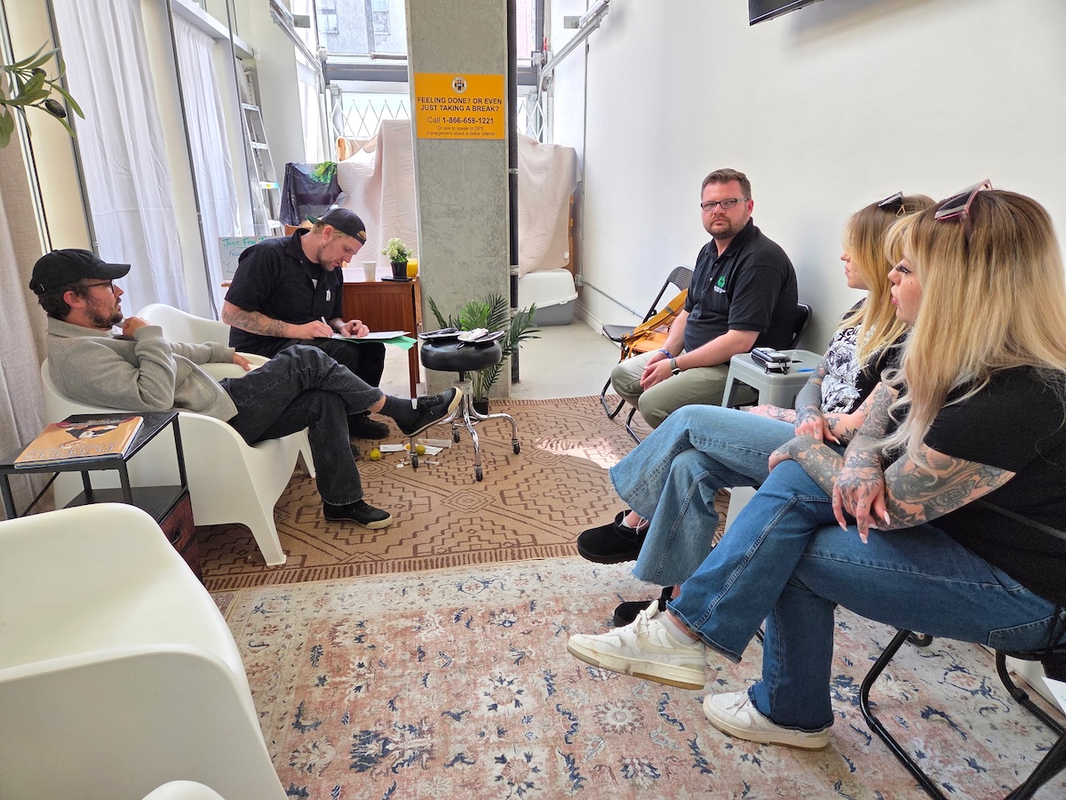 Five people sit on chairs in a semicircle. Trey Helten is taking notes. There are harm reduction supplies at his feet.