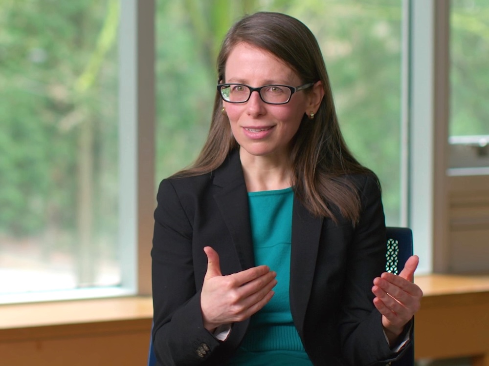 A young to middle-aged white woman with long brown hair and glasses wearing a dark blazer gestures as she talks, sitting in a chair with a window behind her.
