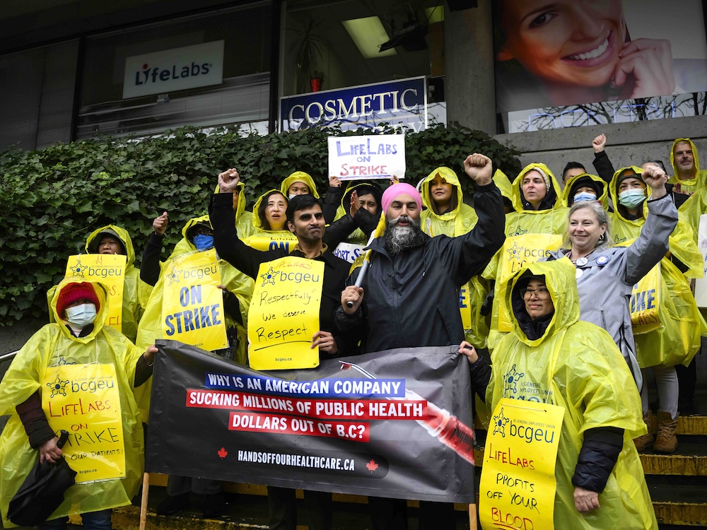 A group of people wearing yellow rain ponchos and signs denouncing LifeLabs stand outside with Jagmeet Singh.