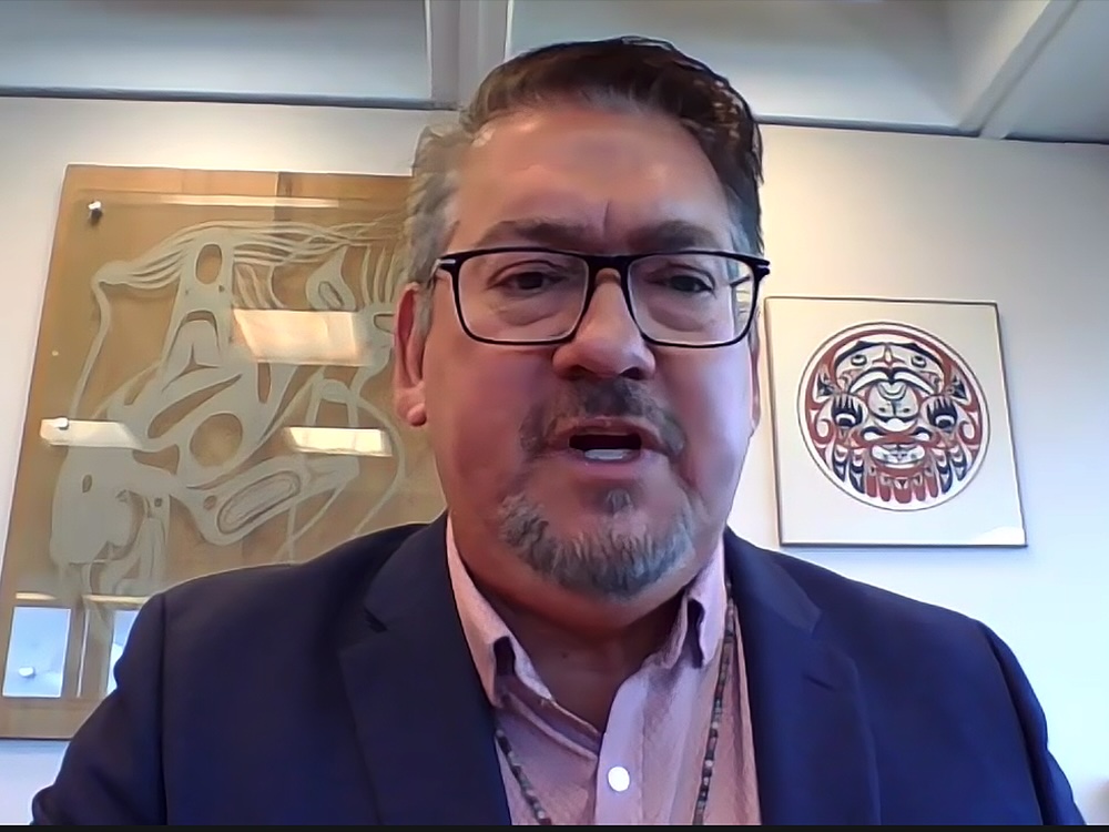A middle-aged man with glasses, short hair and a goatee, and wearing a suit jacket and button-down shirt, speaks on a Zoom call. On the wall behind him are pieces of First Nations art.