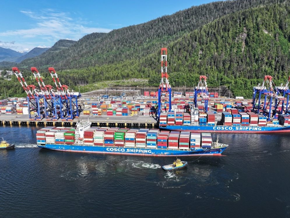 One container ship is docked at a terminal, while tugboats push another one in. The sun is shining and tree-covered hills rise from the port.
