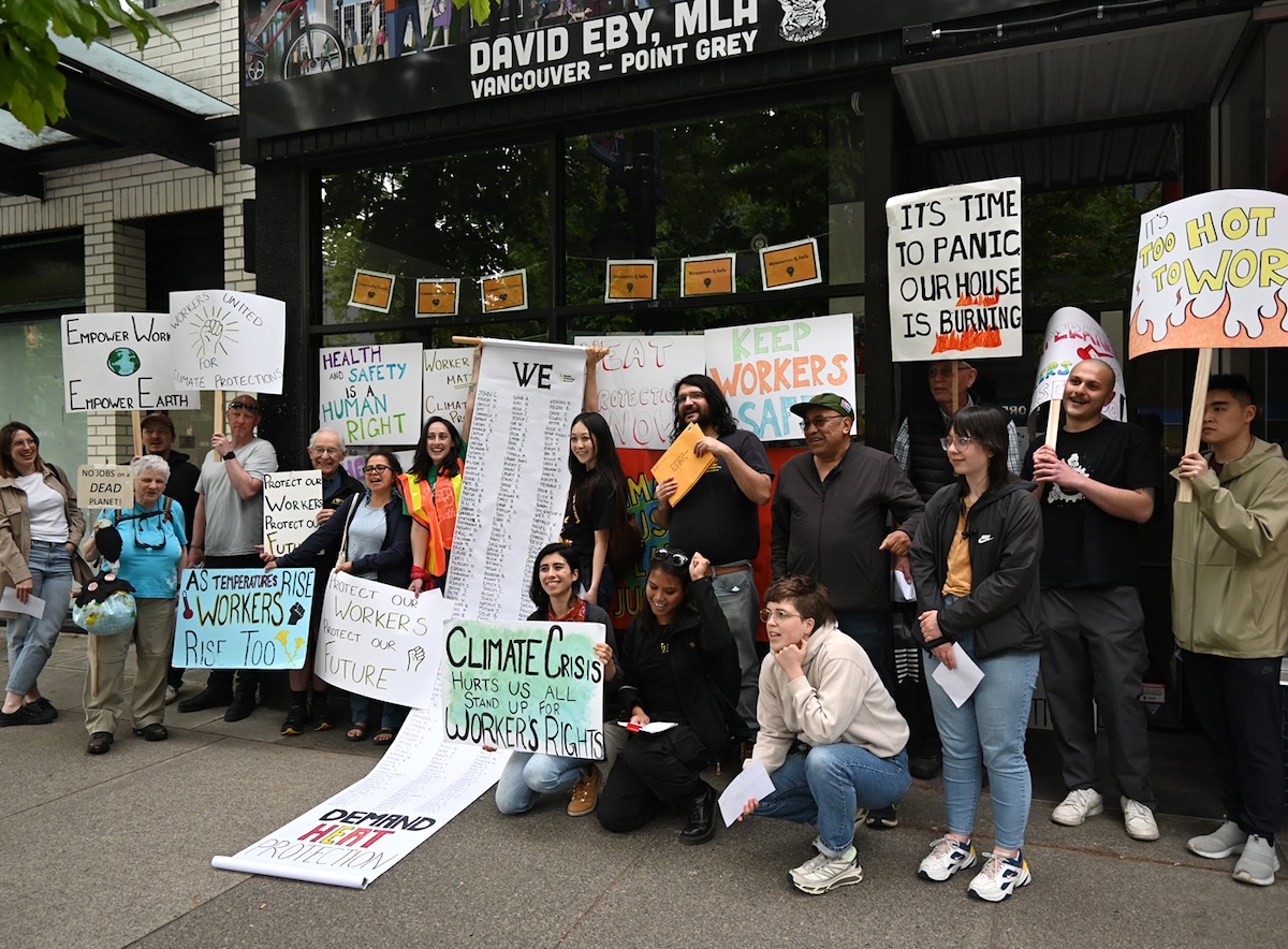 A group of people stand with placards and an unfurled banner outside of David Eby&rsquo;s MLA office. 