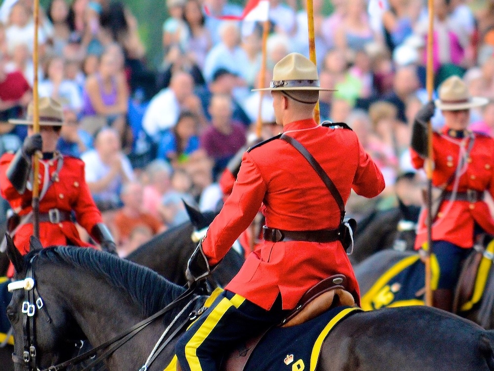 Several Mounties in their serge uniforms ride horses with a crowd out of focus behind them.