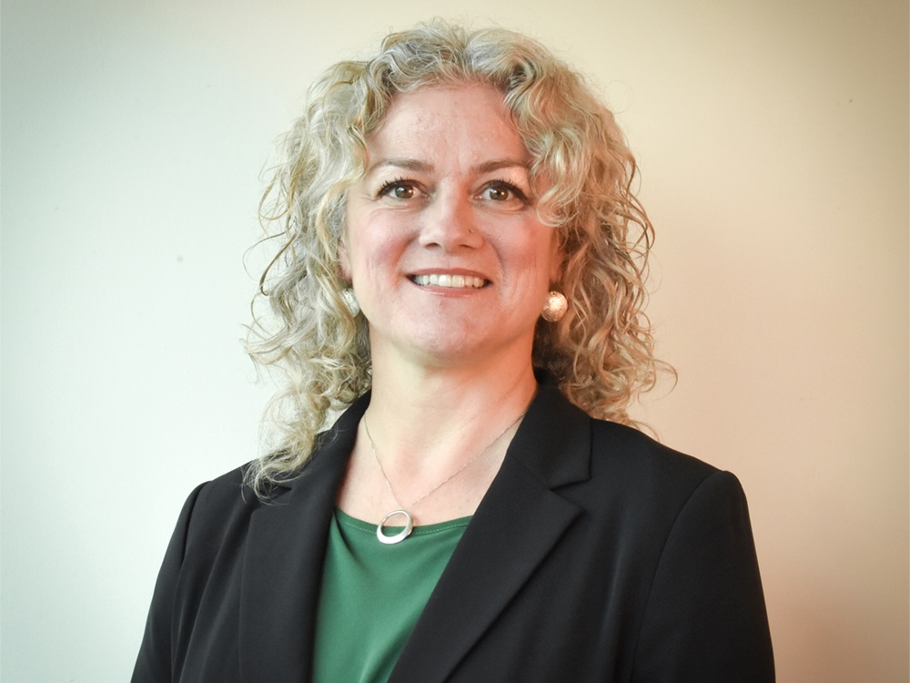 A woman with curly blond-grey hair and light skin tone looks at the camera, wearing a green shirt and a black blazer. 