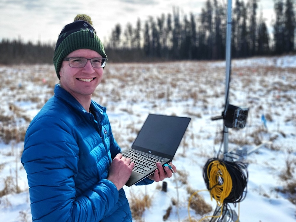 A man with light skin tone wearing glasses, a toque and a blue puffy jacket stands in a snowy field holding a laptop computer.