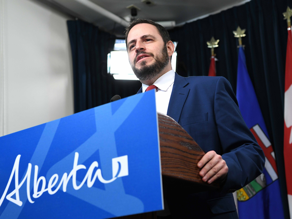 A 42-year-old light-skinned man with dark hair and a trimmed beard stands in a blue suit at a podium.