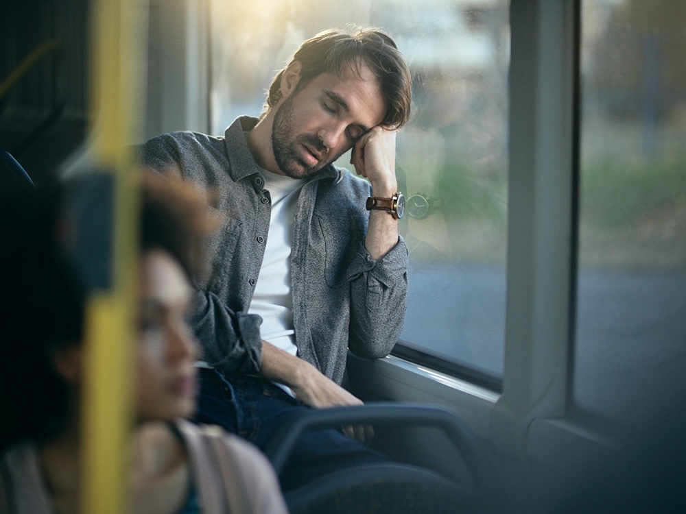 A man with light skin tone and short brown hair leans on a window and sleeps while commuting by bus.