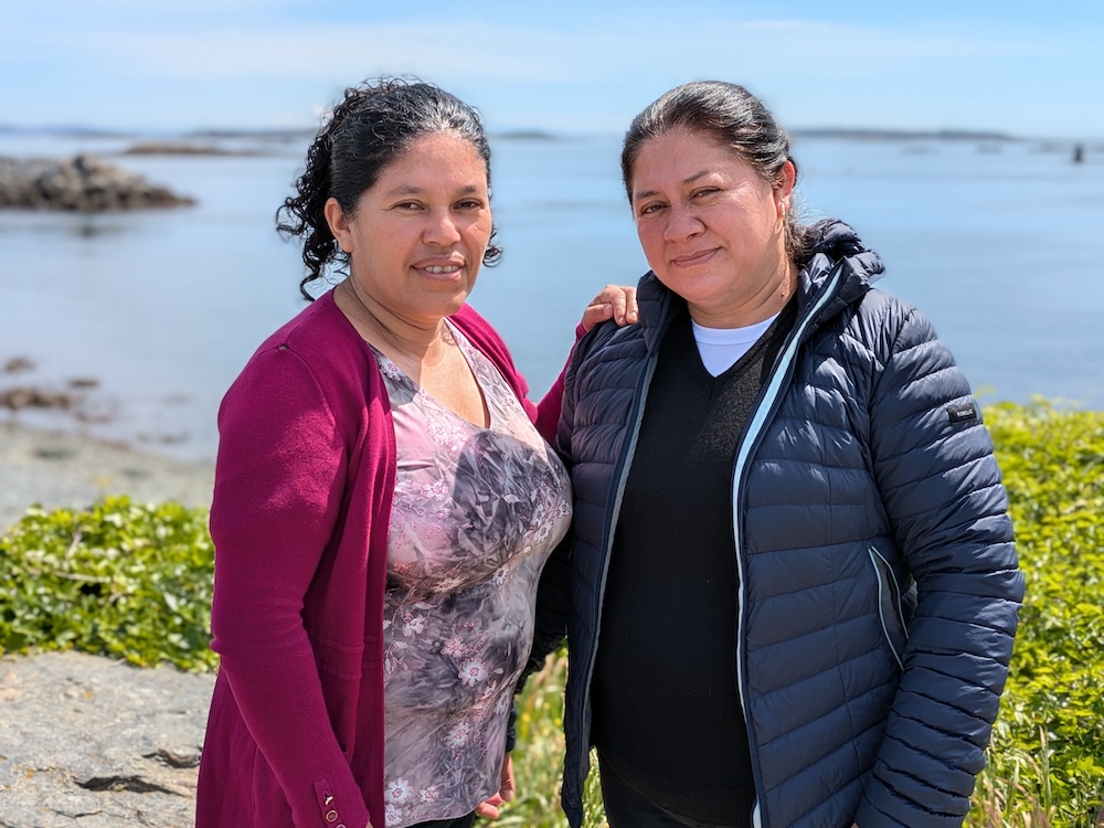 Two women with medium-toned skin and dark hair stand side by side outdoors, with the ocean behind them.