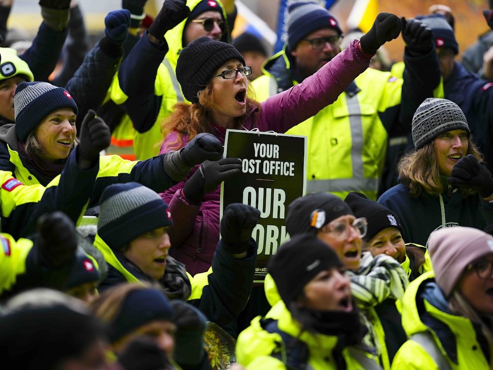 A group of people, some wearing yellow Canada Post jackets, some are raising fists in the air.