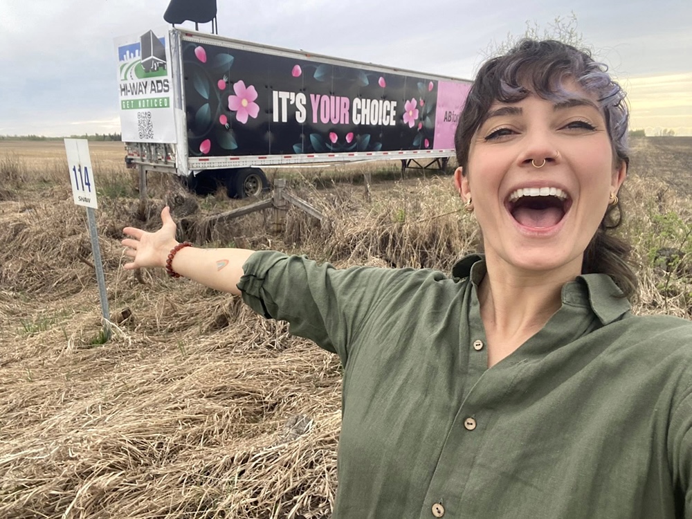 A woman with dark wavy brown and lilac hair and a green shirt stands in front of a white truck parked in a grassy field. Her arm is flung open and she looks overjoyed. &lsquo;It&rsquo;s your choice&rsquo; is written in large pink and white letters on the side of the truck.