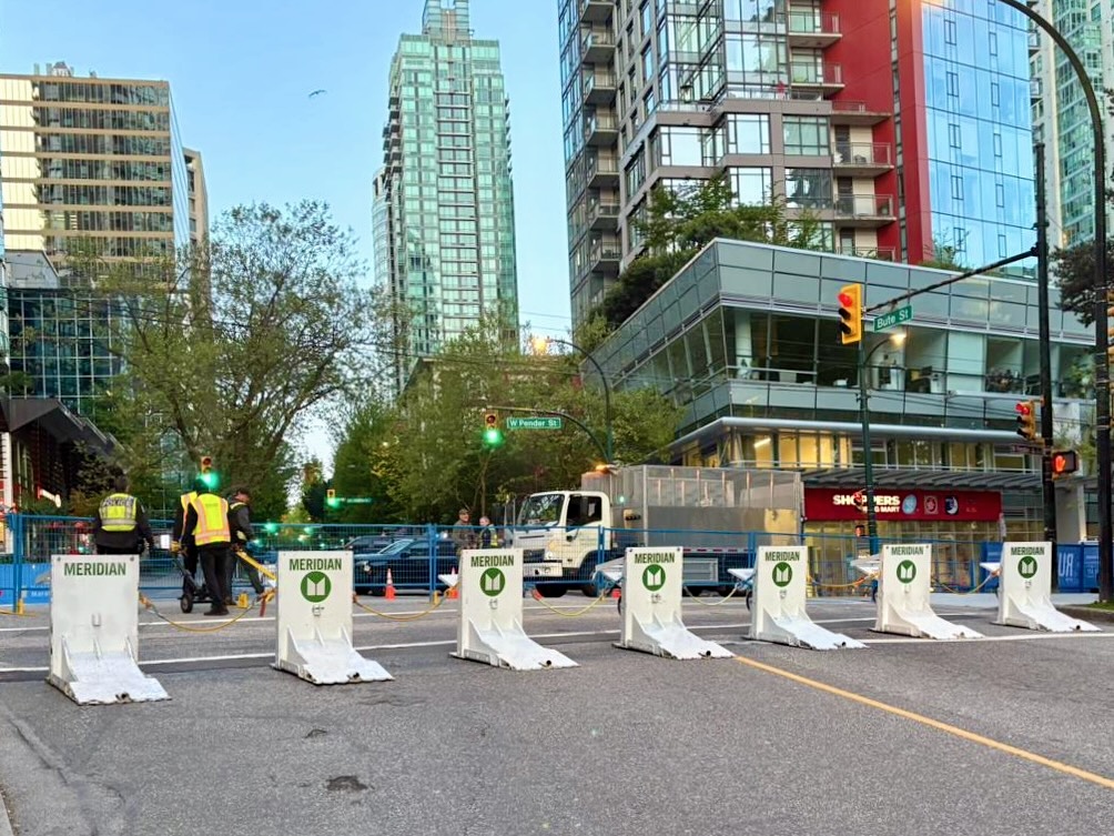 Seven barriers are lined up along a road in front of a metal fence.