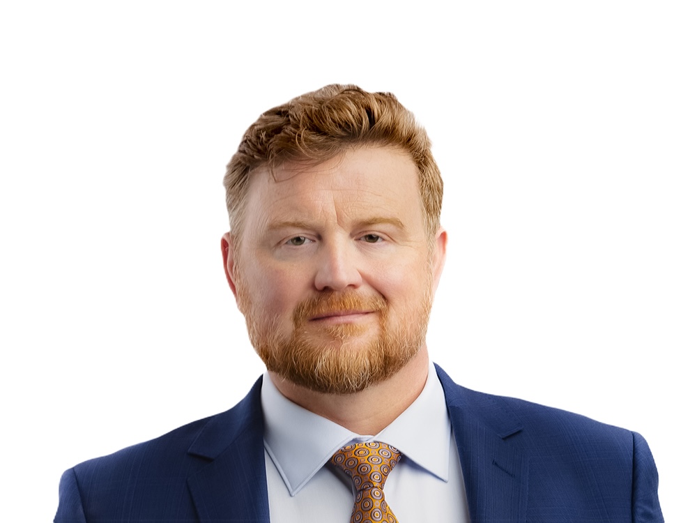 A middle-aged man with red hair and beard, wearing a dark suit and tie, with a white background.