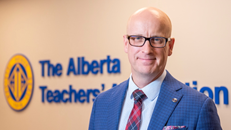 A bald light-skinned man with dark-framed classes in a blue suit stands in front of a wall with the logo and sign of the Alberta Teachers’ Association.