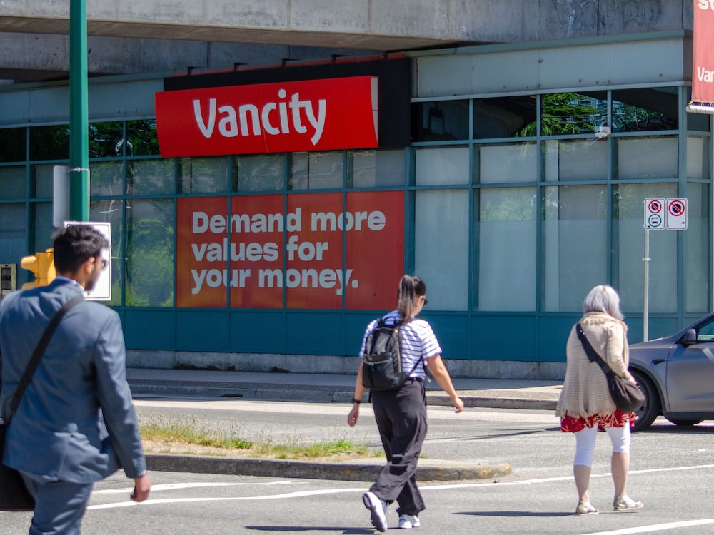 Three people cross a city street by a building with a red and white ‘Vancity’ sign. Another red and white sign below it reads, ‘Demand more values for your money.’