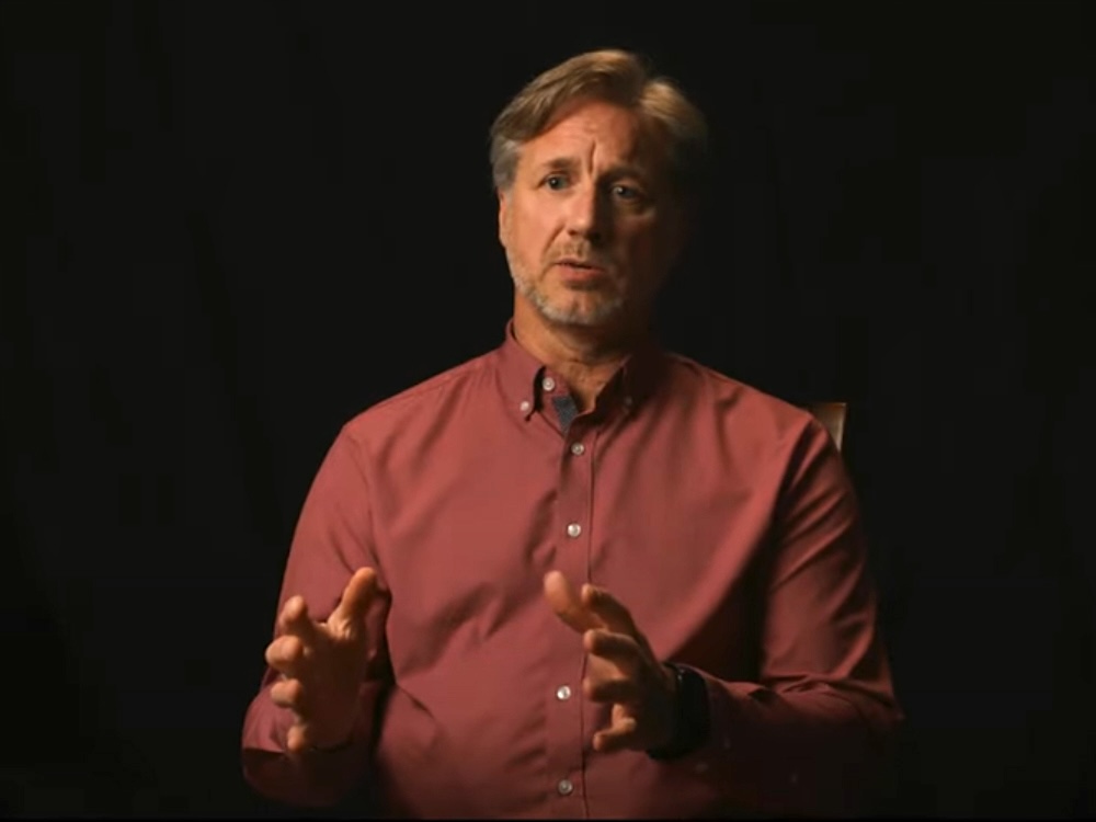Calvin Dubray, a middle-aged man with light skin tone and greying brown hair, speaks to the camera, gesturing with both hands.