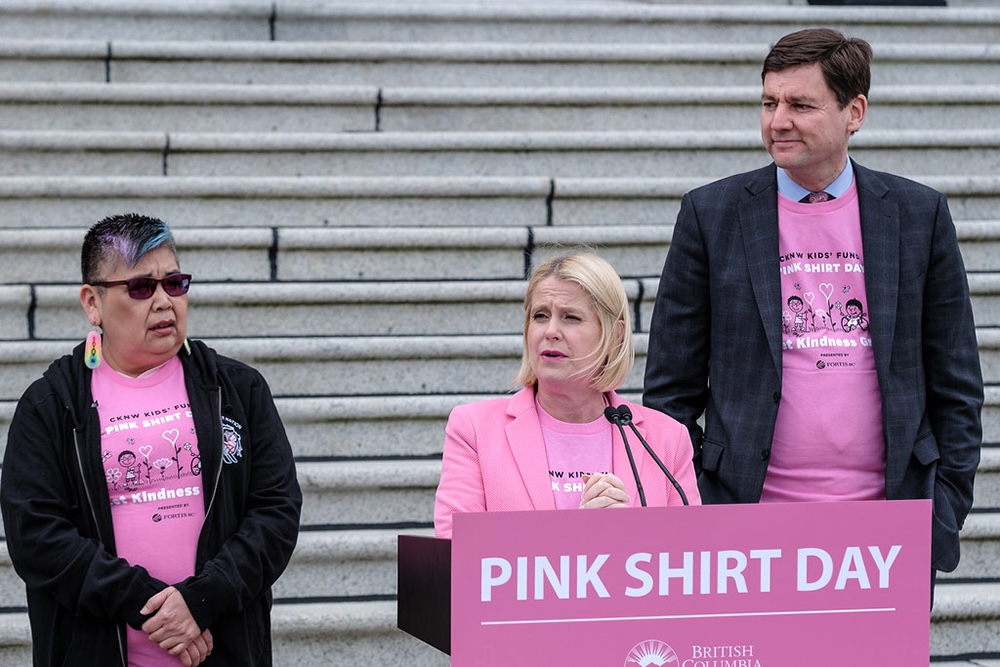 Minister Lisa Beare, a middle-aged woman with light skin tone and blond hair, stands next to Premier David Eby at a ‘Pink Shirt Day’ lectern.