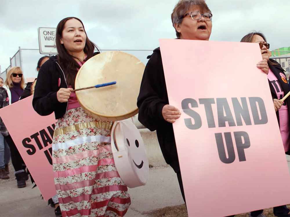 A group of people hold pink signs reading, ‘Stand Up.’ An Indigenous woman wearing a ribbon skirt plays a drum. 