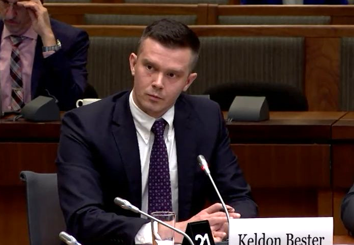 A man with light skin and short brown hair in a dark suit and tie sits in front of a microphone and a sign that reads, &lsquo;Keldon Bester.&rsquo;