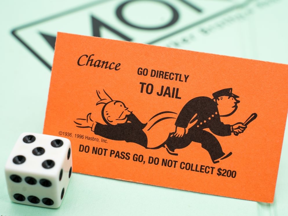 A dice and monopoly playing board with, in the foreground, a &lsquo;Go directly to jail&rsquo; monopoly card showing a rich man being carted off by police.