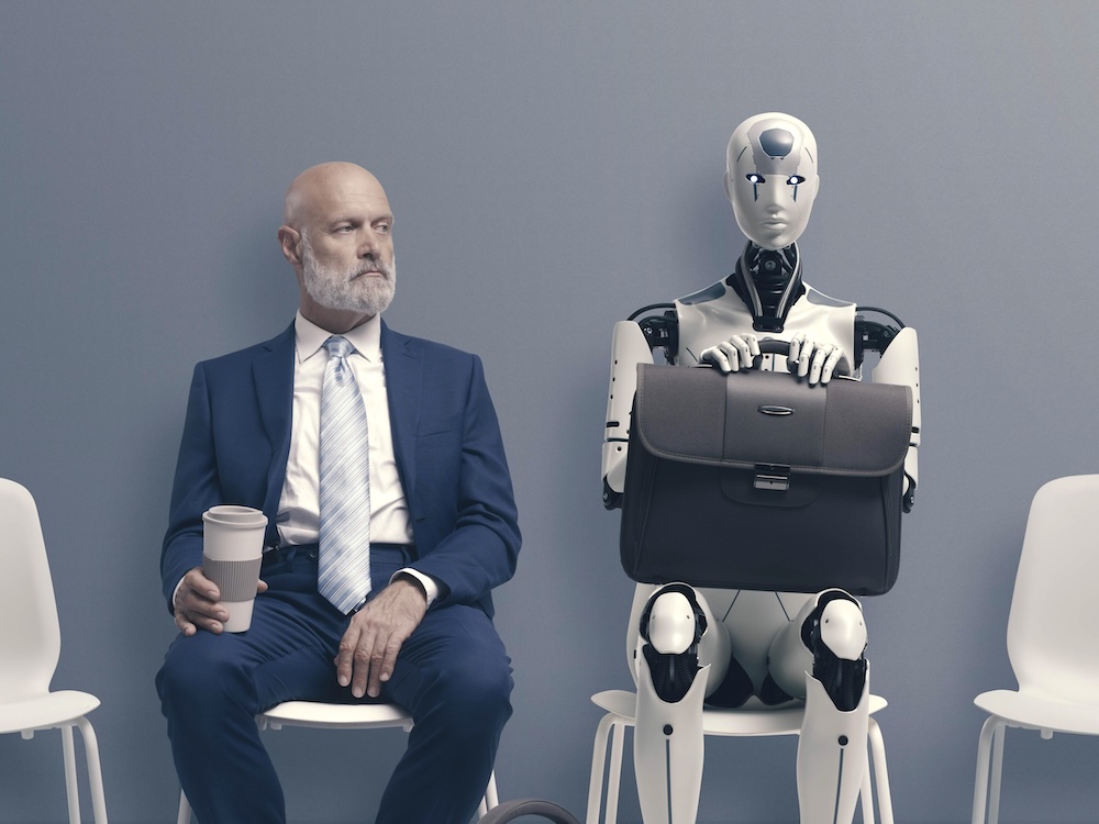 A middle-aged man in a suit sits beside a robot with a briefcase in its lap.