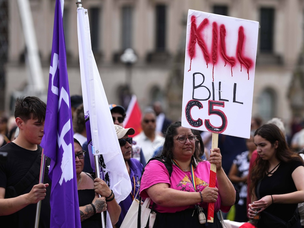 A group of people stand outdoors. One holds a sign that says “Kill Bill C5.”