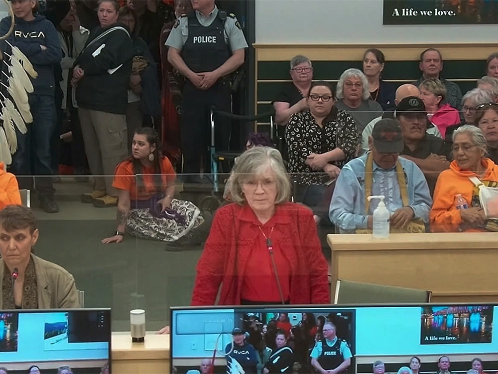 An older woman with light skin tone and a grey bob wears a red collared shirt and red lace blazer and addresses city council. The public audience area appears to be very full behind her, with some people standing and others sitting on the floor.